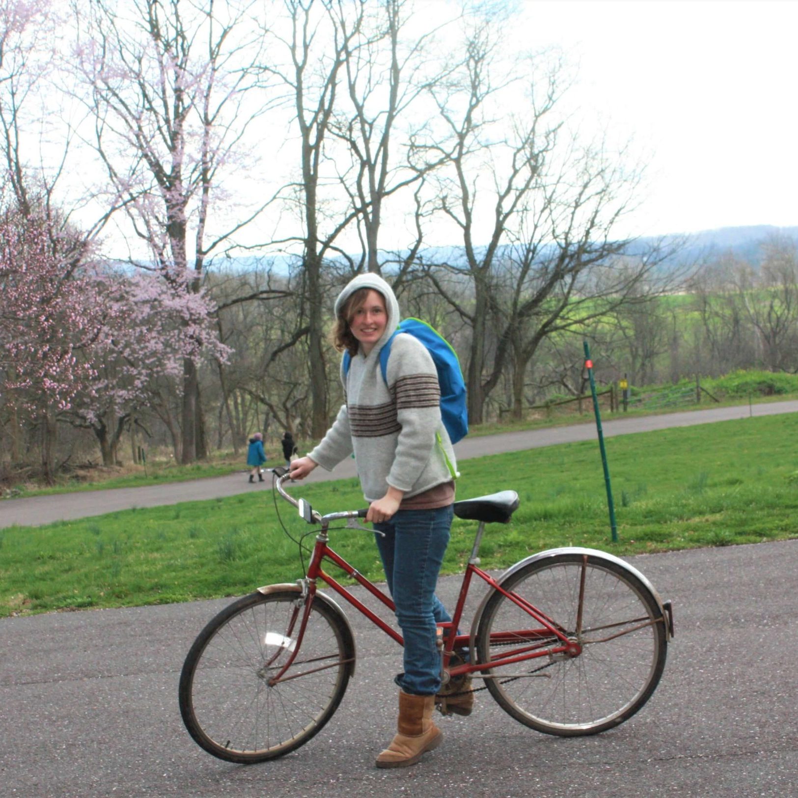 A person wearing a hoodie, winter boots, and a backpack is standing over a bicycle in the road, with a blooming cherry tree, two far away people, and distant hills in the background