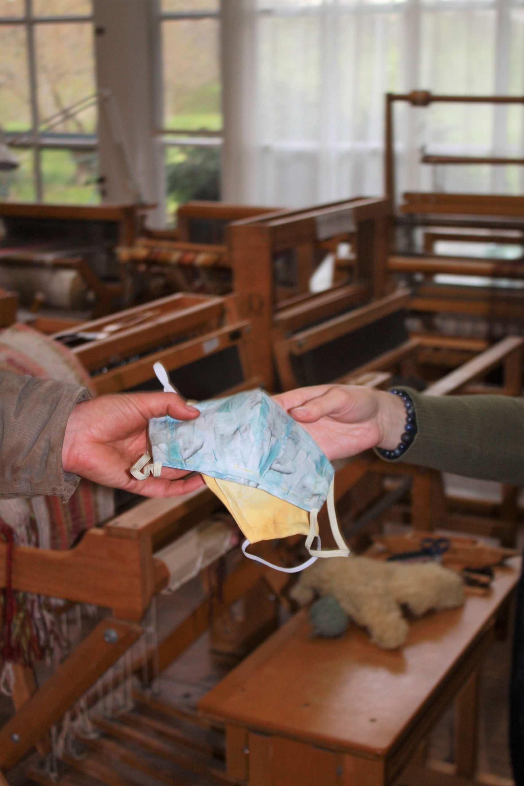 Cloth masks being handed from one hand to another, with looms and big windows in the background