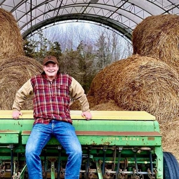 Alex standing on a piece of farm machinery in front of bales of hay, smiling at the camera