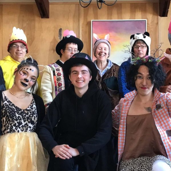 Ike and and nine other Camphilll friends are pictured smiling at the camera in their Halloween costumes. Costumes include a person in a black robe with a mask and cat ears, a person wearing a yellow coat and hat, a person wearing a colorful vest and black hat, a person wearing a brown coat and bunny ears attached to a hat, a person wearing a panda hat, a person wearing a pink dress with a brown jacket and a pink hat shaped like the head of an animal, a person wearing a headband with animal-like ears attached to it, a person wearing a cheetah print shirt with a yellow tutu and cat ears and wearing black lipstick and spotted makeup on their face, Ike wearing a black top hat and a black coat, and a person wearing a dotted skirt with a red checkered shirt and a headband with sticks attached to it