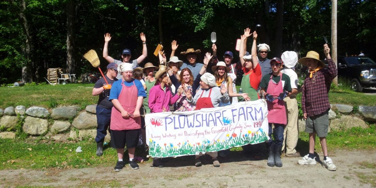 A crowd of people smiling and holding a banner that reads Plowshare Farm Living, Working, and Diversifying the Greenfield Community since 1990