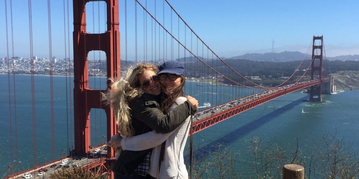 Anna and a friend hugging and smiling at the camera in front of the Golden Gate Bridge