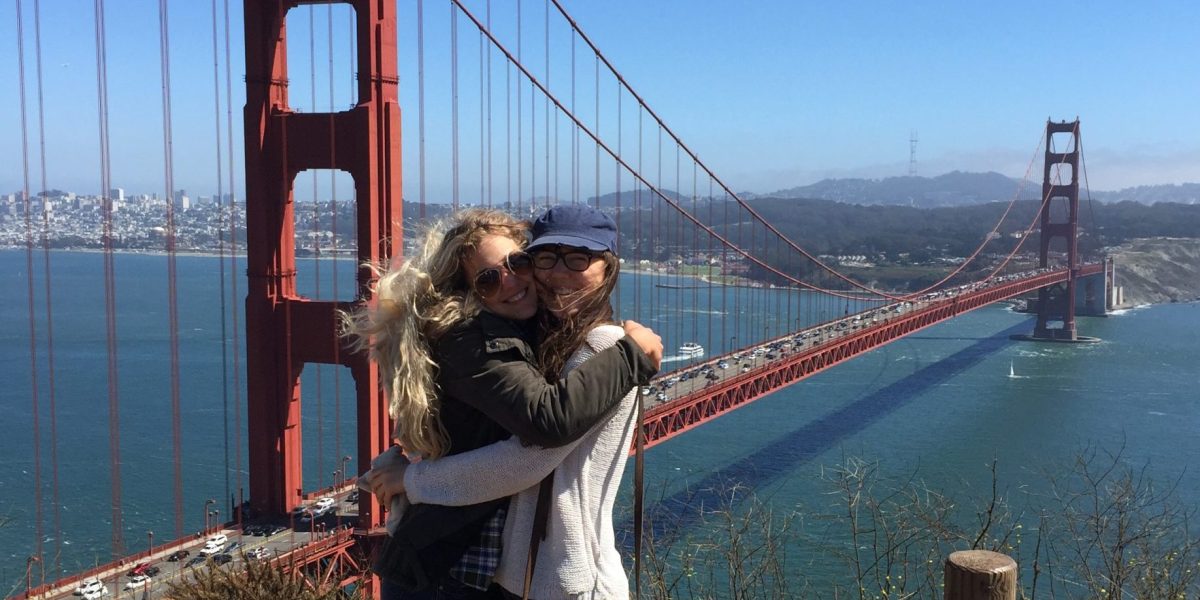 Anna and a friend hugging and smiling at the camera in front of the Golden Gate Bridge