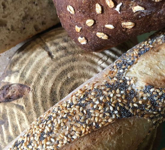 Closeup image of four loaves of bread. One is a round light brown loaf. One is a long loaf with poppy and sesame seeds on it. One is a dark brown loaf with oats on it. The fourth is a slice of a light brown bread. Ike shared this photo of loaves of bread he ate at Camphill, saying "I miss Camphill bread everyday."