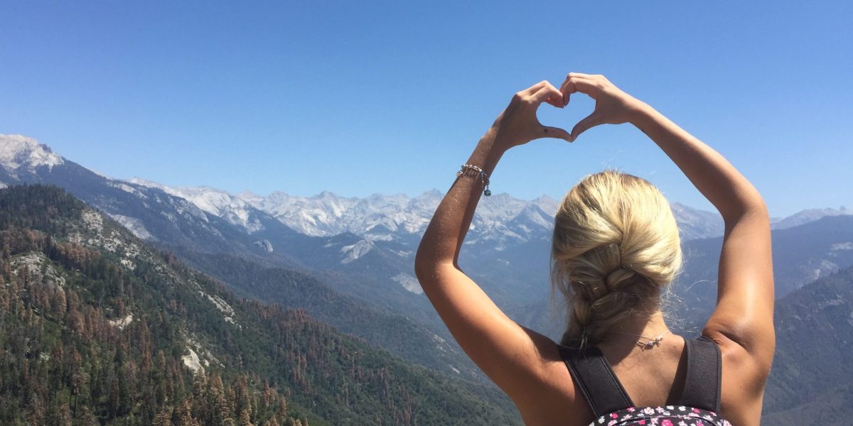 Anna holding her hands in the shape of a heart with her back to the camera with a mountain range in the background