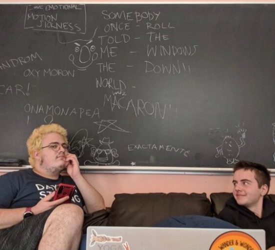 Ike and a friend are sitting on a couch. Ike is looking at his friend, and the friend is looking at the camera while holding a red cell phone. Behind them is a blackboard with small characters and song lyrics drawn on it. The blackboard says "I have emotional motion sickness somebody roll the windows down," "somebody once told me the world wuz macaroni," "onamonapea," "oxymoron," and "exactamente"