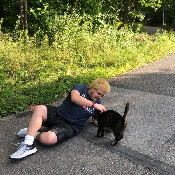 Asher is pictured lying on their side on the concrete while petting a cat. Behind them, there is tall green grass.