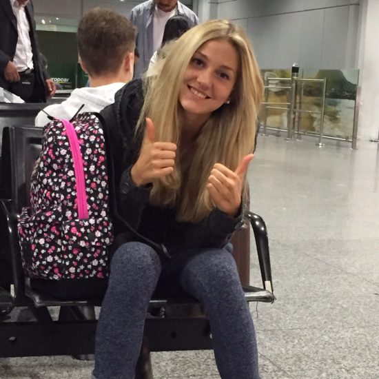 Anna is pictured smiling and holding two thumbs up in the airport on the way to Plowshare Farm