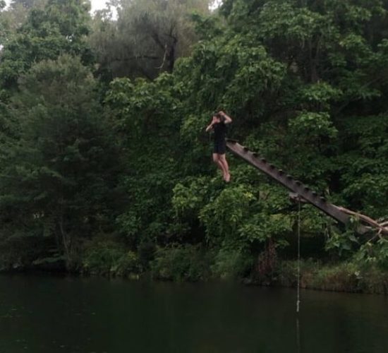 Ike is midair, plugging his with his hand before he lands in the body of water that he is jumping into to go swimming. Behind him are many trees and a tilted wooden ladder that he's jumped off of.