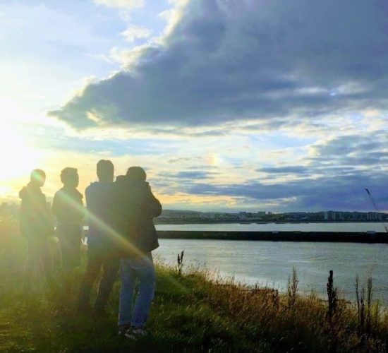 With their backs to the camera, Brittany and three friends look out at the ocean with some buildings and shore in the distance. They are standing in tall grass right by the shore. The sky is slightly cloudy around sunset and a large beam of sunlight covers much of the photograph.