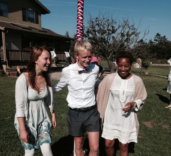 Brittany smiles at the camera with two friends. They are all wearing white clothing and are standing in front of a may pole wrapped in ribbon. There is a brown building behind them and a bright blue sky in the background.