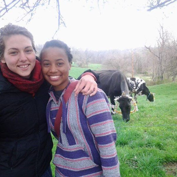 Brittany, wearing a pink and purple striped hoodie smiles with her arm around a friend who is also smiling while wearing a black coat and a red scarf. Behind them are two horses in a large field of trees and grass.