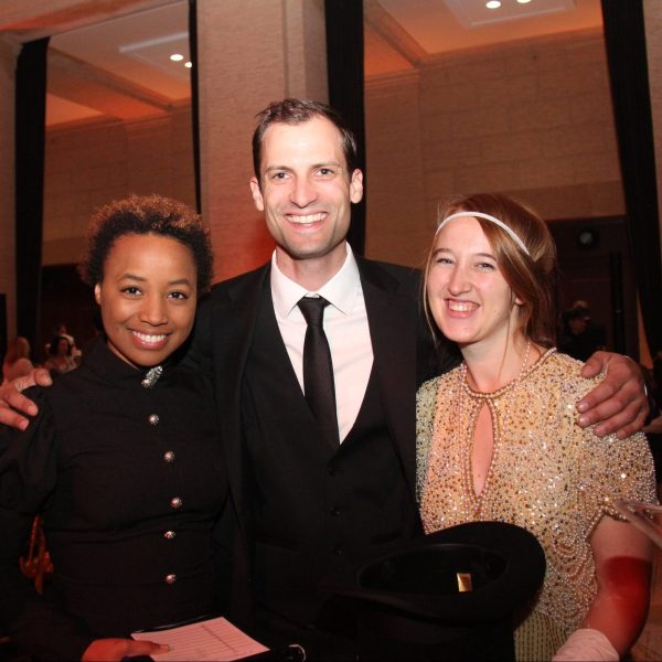 Brittany, left, is smiling in a fancy, black button up dress with a brooch at her neck. Next to her is a person in suit and tie and another person wearing a gold sequin dress and white gloves. They are all smiling, and behind them there are nicely set tables with candles, flowers, and golden chairs.