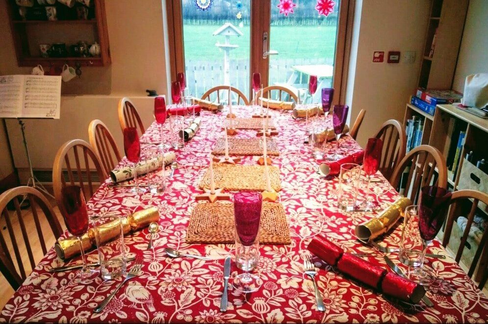 A table with ten chairs around it and a beautiful red table cloth with white birds and flowers on the fabric. The table has champagne flutes, silverware, water cups, candles, placemats, and shiny wrapped at each chair.. In the background there is a music stand, a rack of mugs, a cabinet filled with books and puzzles, and a door to the outside with decorations on it. Outside, there is a stand for birdseed and a picnic table.