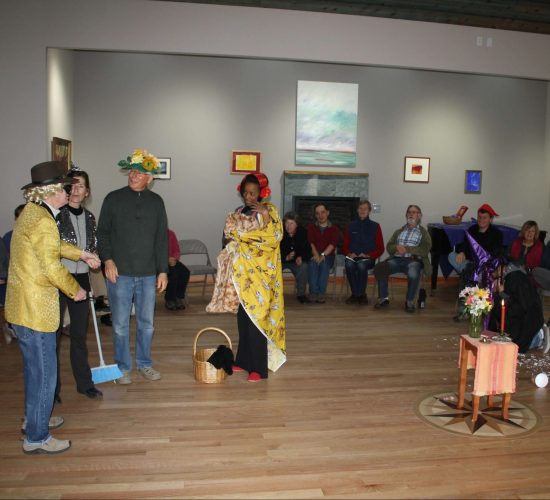 Brittany and three friends stand in the middle of a large circle of Camphill villagers and volunteers. They are performing in a play. One of them wears a yellow jacket and a brown top hat, one wears a sequin jacket and holds a broom, one wears a green hat covered in flowers, and Brittany wears a red hat and is wrapped in a yellow blanket while holding a basket with a stuffed dog in it. She is playing Dorothy in the Wizard of Oz.