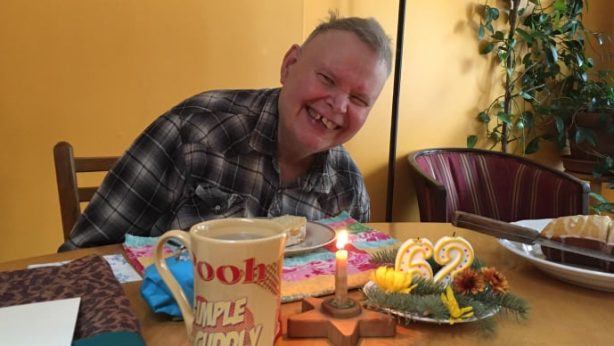 Jeff is wearing a black and white plaid shirt. He is sitting at a table, and in front of him is a mug, a slice of birthday cake, the rest of the birthday cake, a lit candle, and two unlit candles that say 62. He is smiling.