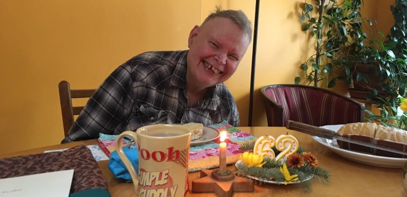 Jeff is wearing a black and white plaid shirt. He is sitting at a table, and in front of him is a mug, a slice of birthday cake, the rest of the birthday cake, a lit candle, and two unlit candles that say 62. He is smiling.