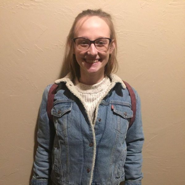 Anna is wearing a white sweater with a jean jacket over it and carrying a backpack on her back. She is standing against a wall, smiling.