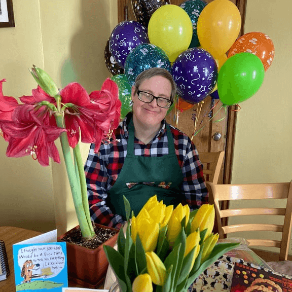 Ben is wearing a red and black plaid shirt with a green apron over it. He is sitting at a table. On the table in front of him are birthday cards, a package of cookies, a plant with pink flowers on it, and a plant with yellow flowers on it. Behind him are many balloons. He is smiling.