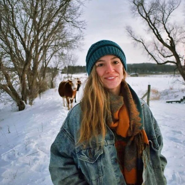 Charlotte is wearing a jean jacket, an orange and green scarf, and a blue beanie. She is standing on a snowy path with trees on either side of her, and behind her there is a brown and white cow. Charlotte is smiling.