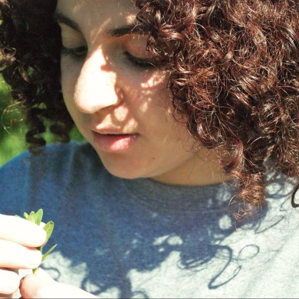 Clara is wearing a grey t-shirt and is looking down at a small green plant that she's holding.