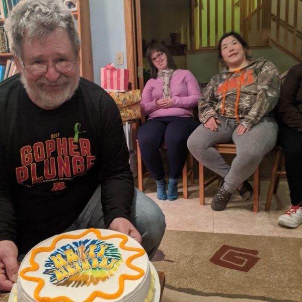 Ben is wearing a black tshirt that says "gopher plunge." He is holding a cake decorated with yellow, orange, and white icing that says "happy birthday on it." He is smiling. Two friends are smiling in the background.