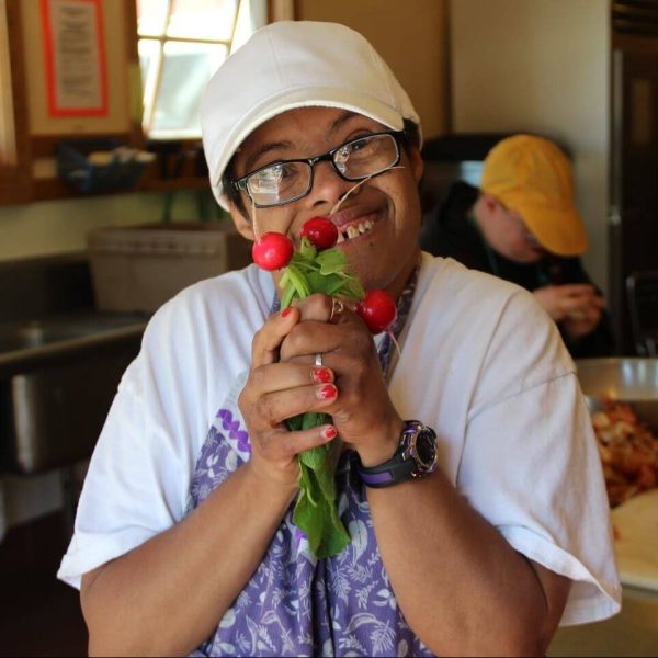 Dede is wearing a white tshirt, a white baseball cap, and a purple apron. She is smiling while holding two handfuls of radishes in front of her face.
