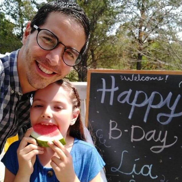 Diego is wearing a blue and white plaid shirt. He is smiling and has his arm around Lia, who is wearing a blue shirt. She is eating a slice of watermelon. Behind them is a sign that says "Welcome! Happy B-Day Lia and Diego"