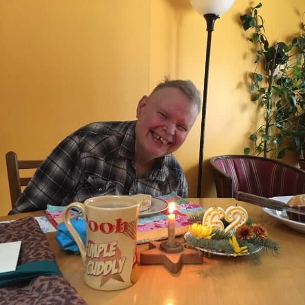 Jeff is wearing a black and white plaid shirt. He is sitting at a table, and in front of him is a mug, a slice of birthday cake, the rest of the birthday cake, a lit candle, and two unlit candles that say 62. He is smiling.