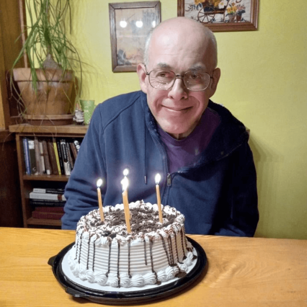 John is wearing a blue zipped hoodie over a purple tshirt. In front of him is a birthday cake decorated with white icing and chocolate candy. He is smiling.