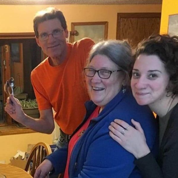 Kristin is wearing a red tshirt with a blue jacket over it. She is smiling and serving ice cream. She is being hugged from behind by another smiling person wearing a black shirt, and another smiling person in an orange shirt is holding spoons behind them.