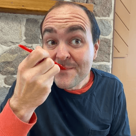 Lee is wearing an orange and black tshirt, and he is smiling with a spoon in his mouth.