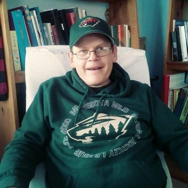 Mike is wearing a green Minnesota Wild NHL hoodie and a green team hat as well. He is sitting in a chair in front of bookcases and smiling.