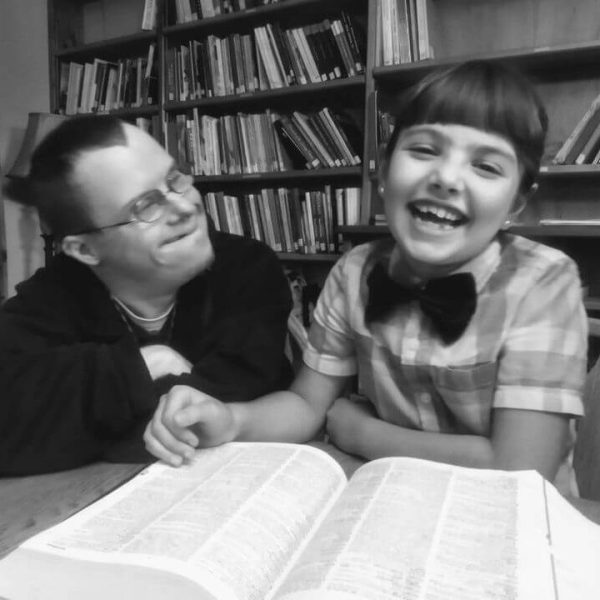 Monte is wearing a black shirt. He is smiling while looking at a child who is wearing a plaid shirt and bowtie and is smiling at the camera. In front of them is a large book sitting on a table, and several bookcases of books are behind them as well.