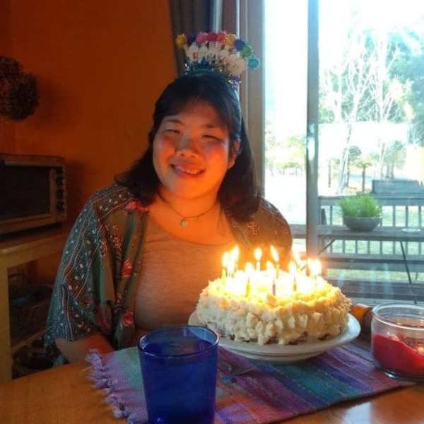 Nae Nae is wearing a grey tshirt and a green and red jacket over it. She also has a pom pom headband that says "happy birthday." She is smiling. In front of her there is a birthday cake with white icing covered in many candles.