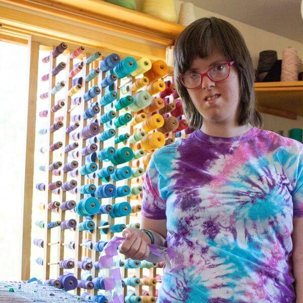 Nicole is wearing a pink, purple, and blue tie dye tshirt. She is holding blue and purple ribbons that she is threading through a weaving loom. Behind her are many other spools of ribbons and threads arranged in a rainbow.