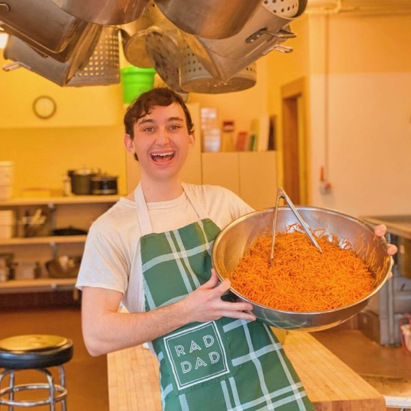 Nicolo smiles for the camera while holding a large metal bowl full of spaghetti. He is wearing an apron that reads "rad dad" and he is standing in a kitchen with many pots hanging from the ceiling above him.