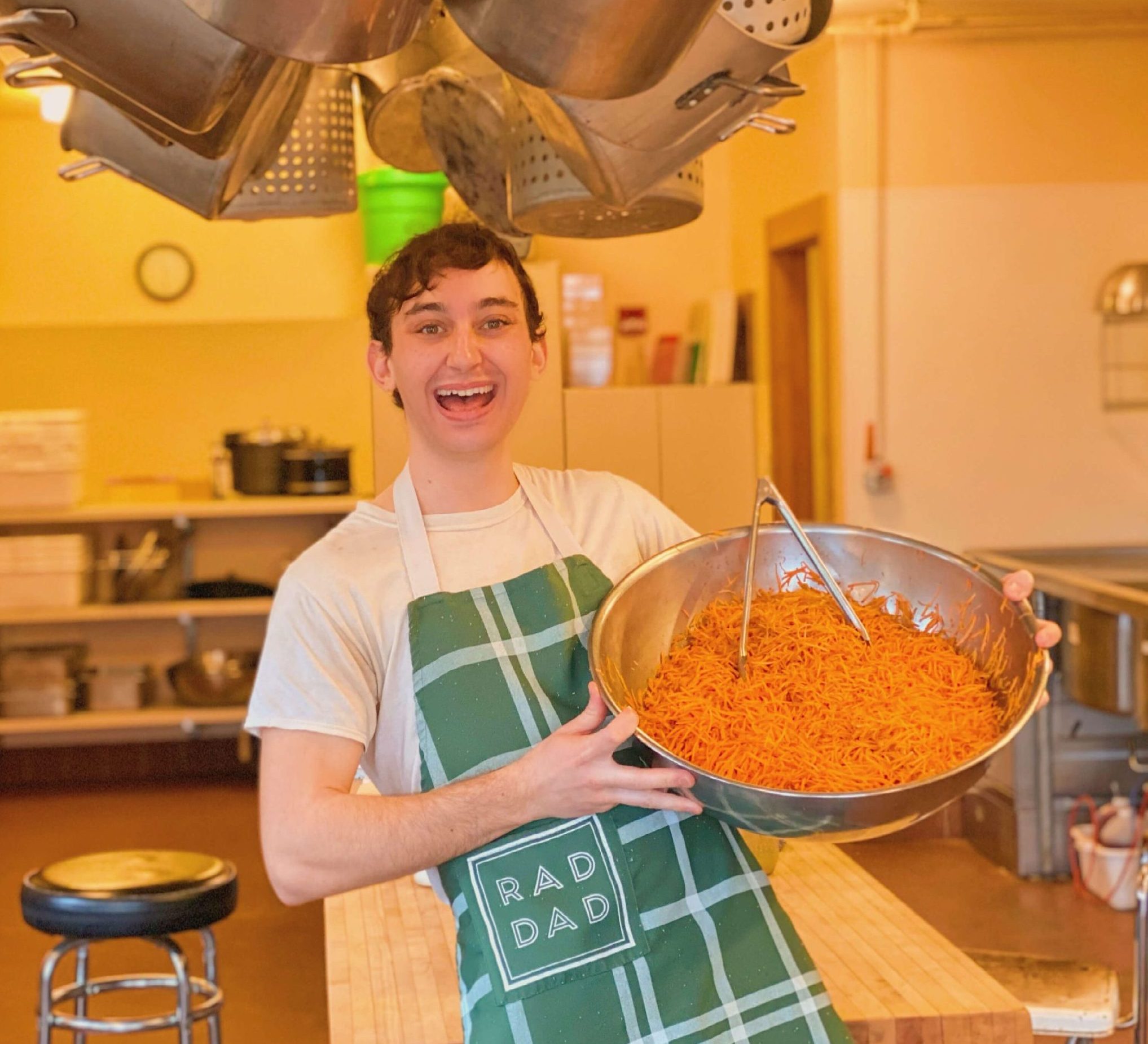 Nicolo smiles for the camera while holding a large metal bowl full of spaghetti. He is wearing an apron that reads "rad dad" and he is standing in a kitchen with many pots hanging from the ceiling above him.
