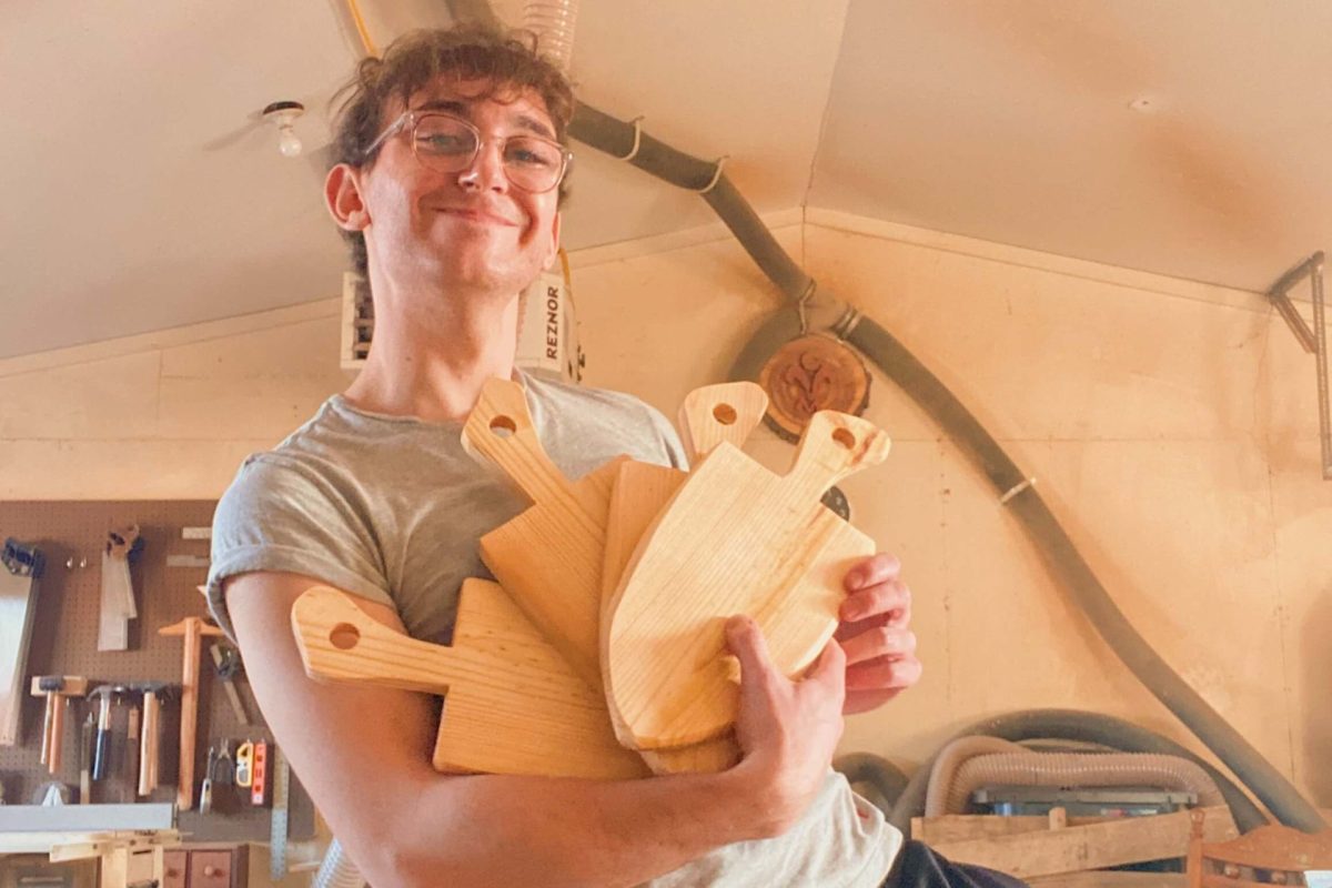 Nicolo smiles while standing in the woodworking shop. He is holding four cutting boards.