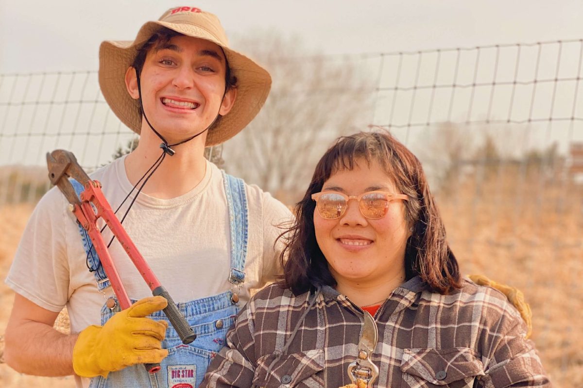 Nicolo and a friend smile while holding large hedge clippers. They are standing outside on the farm, and Nicolo is wearing a large sun hat and overalls. His friend is wearing a plaid brown jacket and sunglasses.