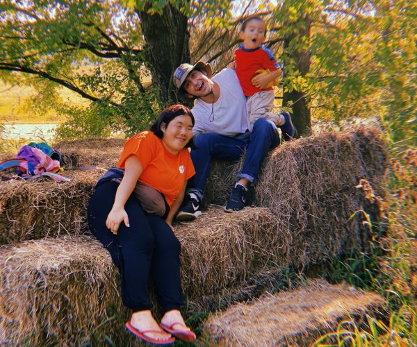 Nicolo and two friends smile for the camera while sitting on a pile of hay. Behind them are many trees and a body of water.