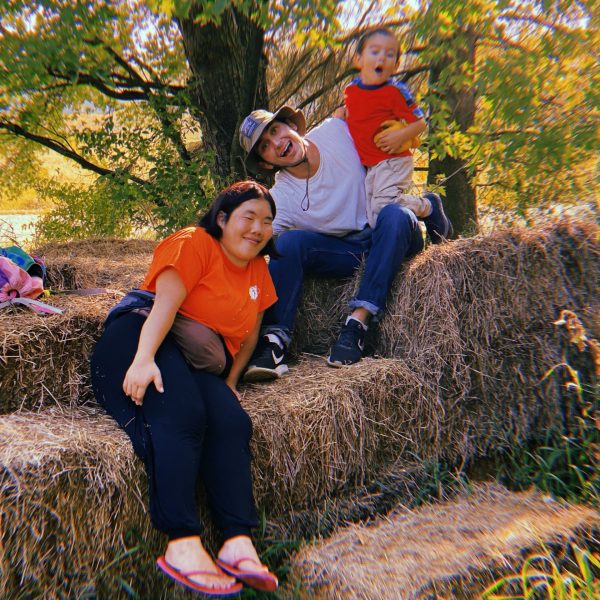Nicolo and two friends smile for the camera while sitting on a pile of hay. Behind them are many trees and a body of water.