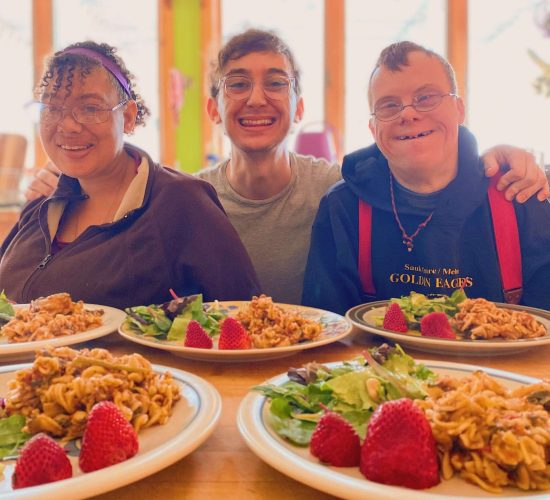Nicolo sits between two friends at a table, all three are smiling, and Nicolos arms are around the friends. In front of them are five plates of food.