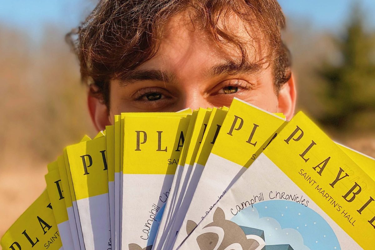 Closeup picture of Nicolo holding a large stack of playbills fanned in front of his face, with only his eyes and above showing. The visible part of the playbills shows a mountain with trees and animals in front of it and the words "Camphill Chronicles."