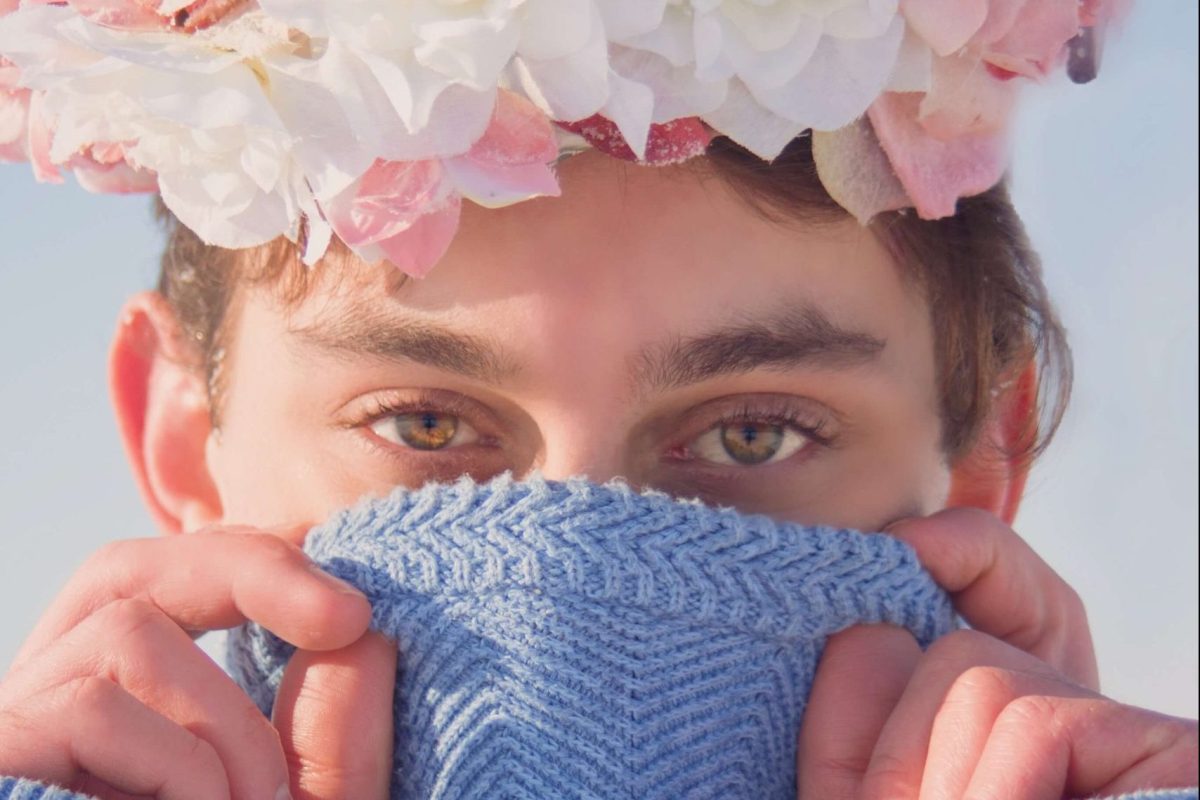 Nicolo is wearing a blue sweater and a crown of pink and white flowers. He is holding the sweater up over his nose and mouth and looking into the close-up camera with sunlight on half of his face.