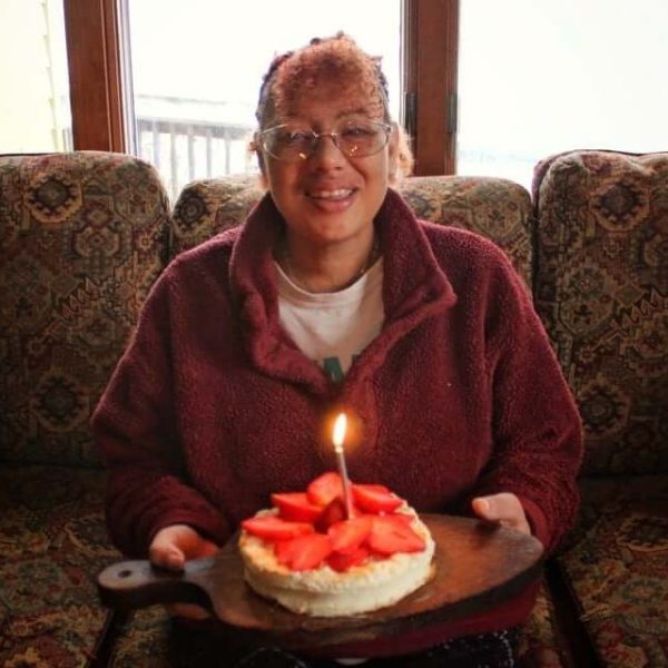 Sara is wearing a red sweatshirt over a white tshirt. She is smiling while holding a cake with white icing, strawberries, and a single lit candle.