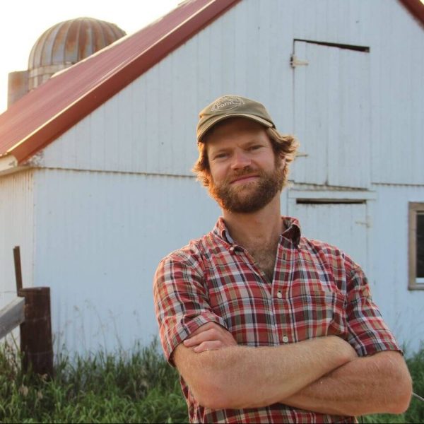 Stephen is wearing a red plaid shirt and a light brown baseball cap. He is crossing his arms and looking at the camera while standing in front of a barn.
