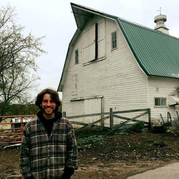 Stuart is wearing a black and brown plaid jacket over a black hoodie. He is smiling at the camera with his hands in his pockets while standing in front of a barn.