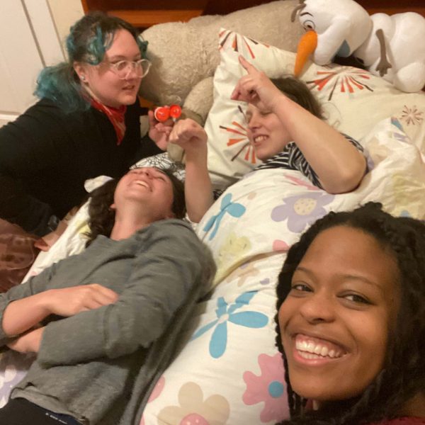 Gabi and three Camphill friends lie in a bed together after having tucked one friend in. Gabi is smiling and taking a selfie of the four of them, and her three friends are all looking at each other and laughing.