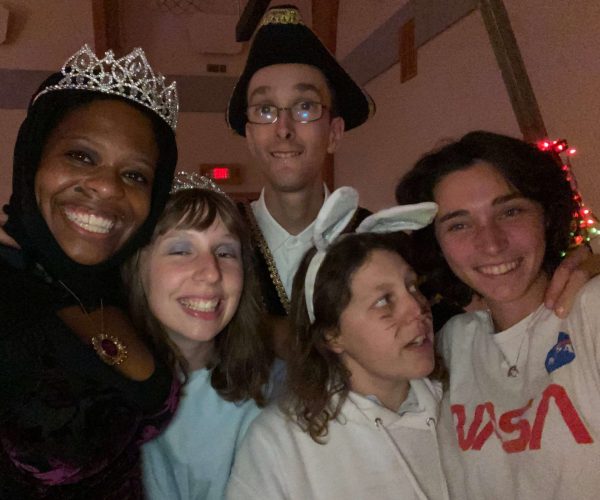 Gabi and four friends smile for the camera while dressed in costumes for Halloween. Gabi is wearing a dark purple dress and a tiara, the second friend from the left is wearing a tiara and a blue dress, the friend in the back is wearing a triangular black hat with gold stitching on it, the second friend from the right is wearing a white hoodie and white bunny ears, and the friend on the right is wearing a NASA tshirt.